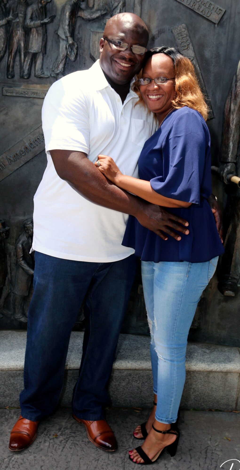 A man and woman smiling and hugging, standing on stairs with a textured wall background.