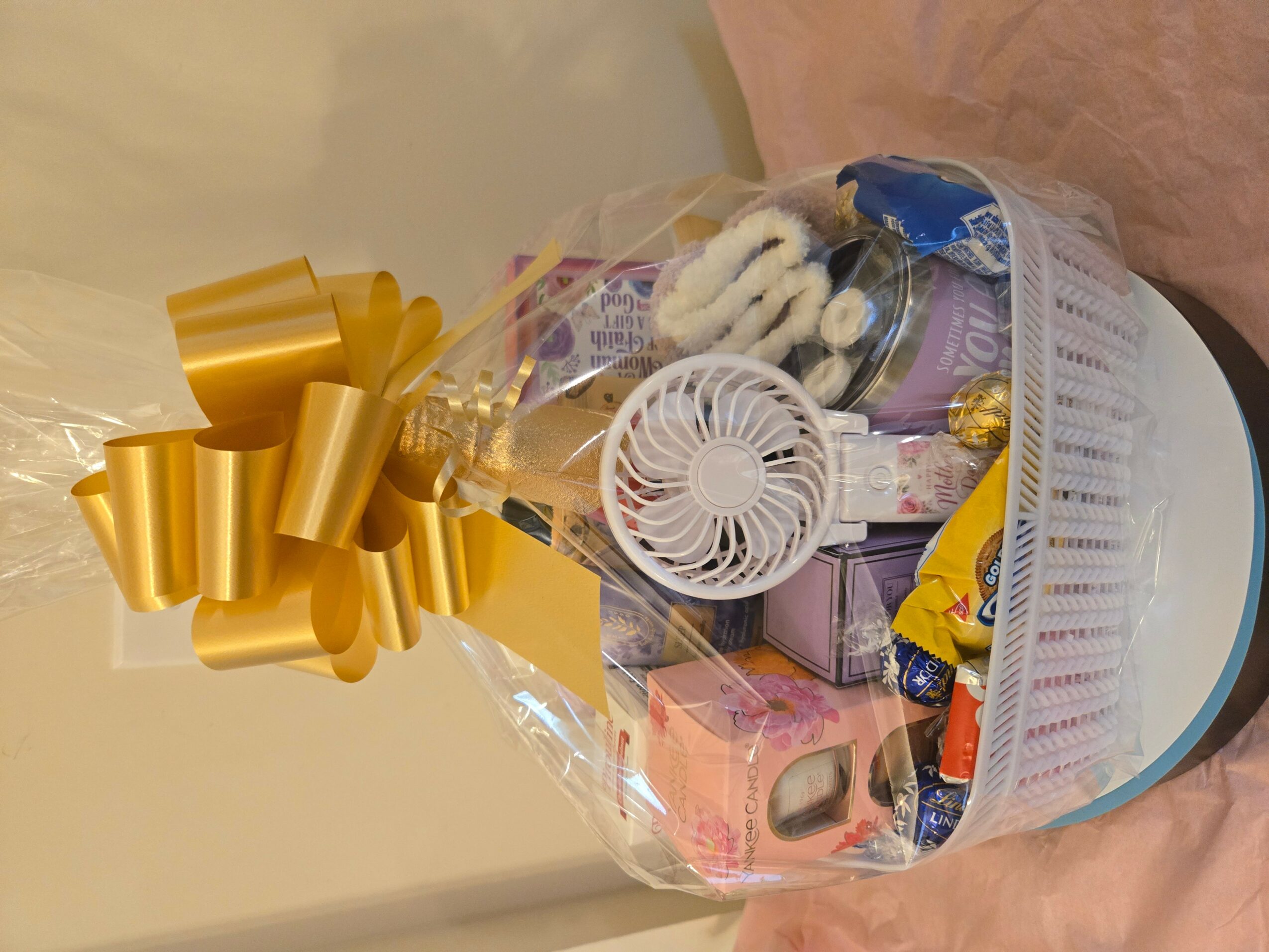 Wrapped gift with a large gold ribbon, containing various small items and a white fan, on a round surface.