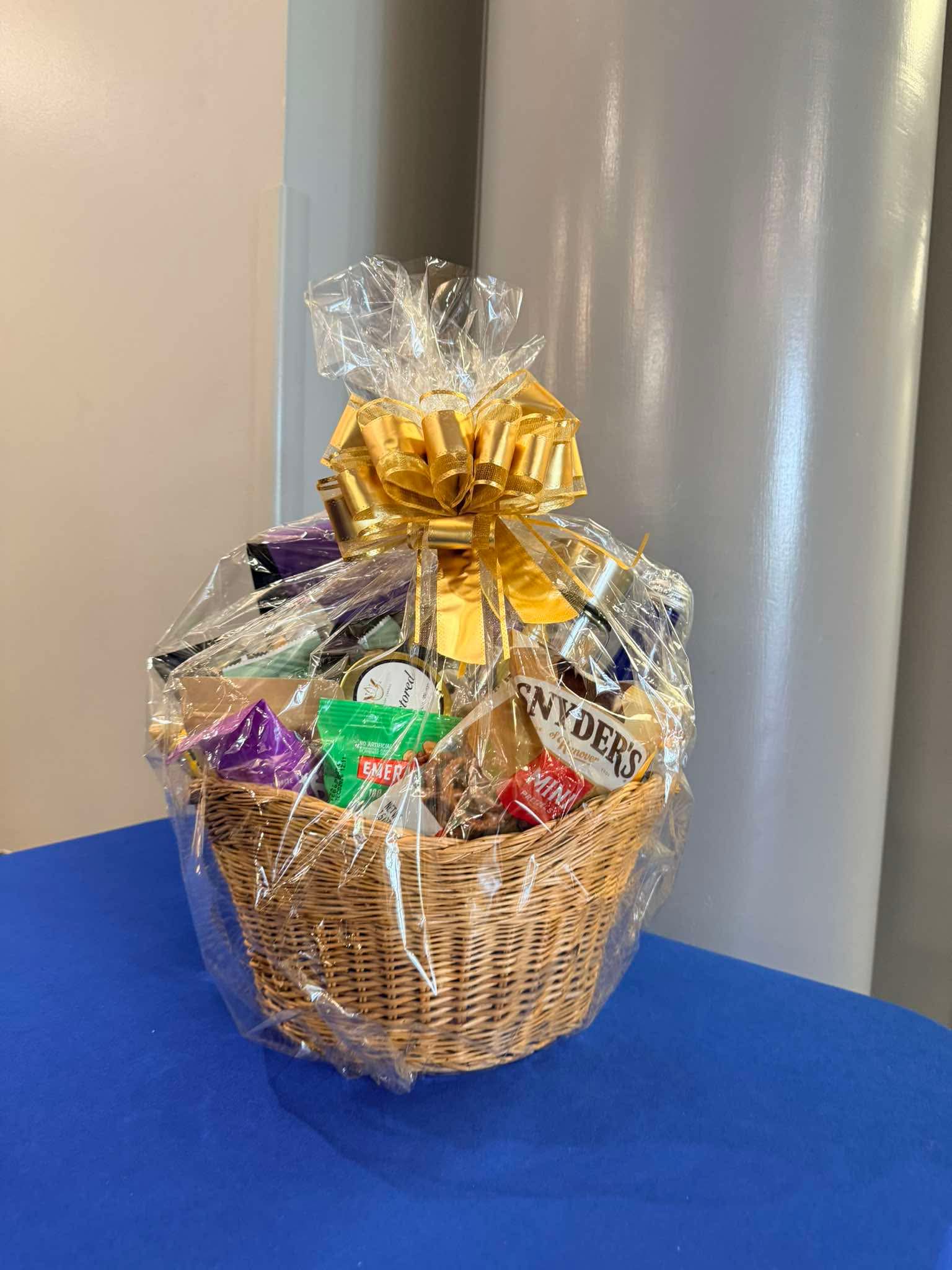 Gift basket wrapped in clear plastic with a large gold bow, containing snacks and treats, on a blue surface.