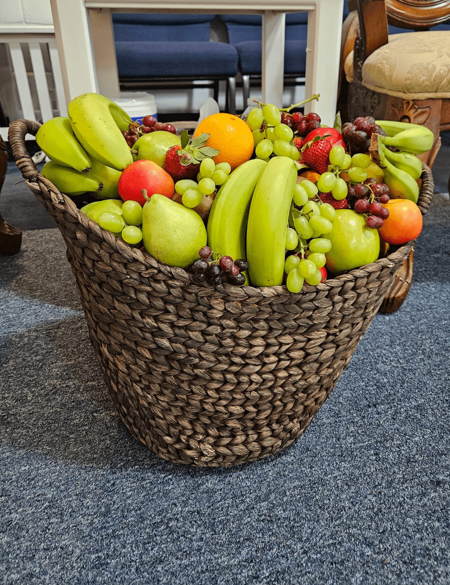 Basket filled with bananas, grapes, apples, oranges, and other fruits on a carpeted floor.
