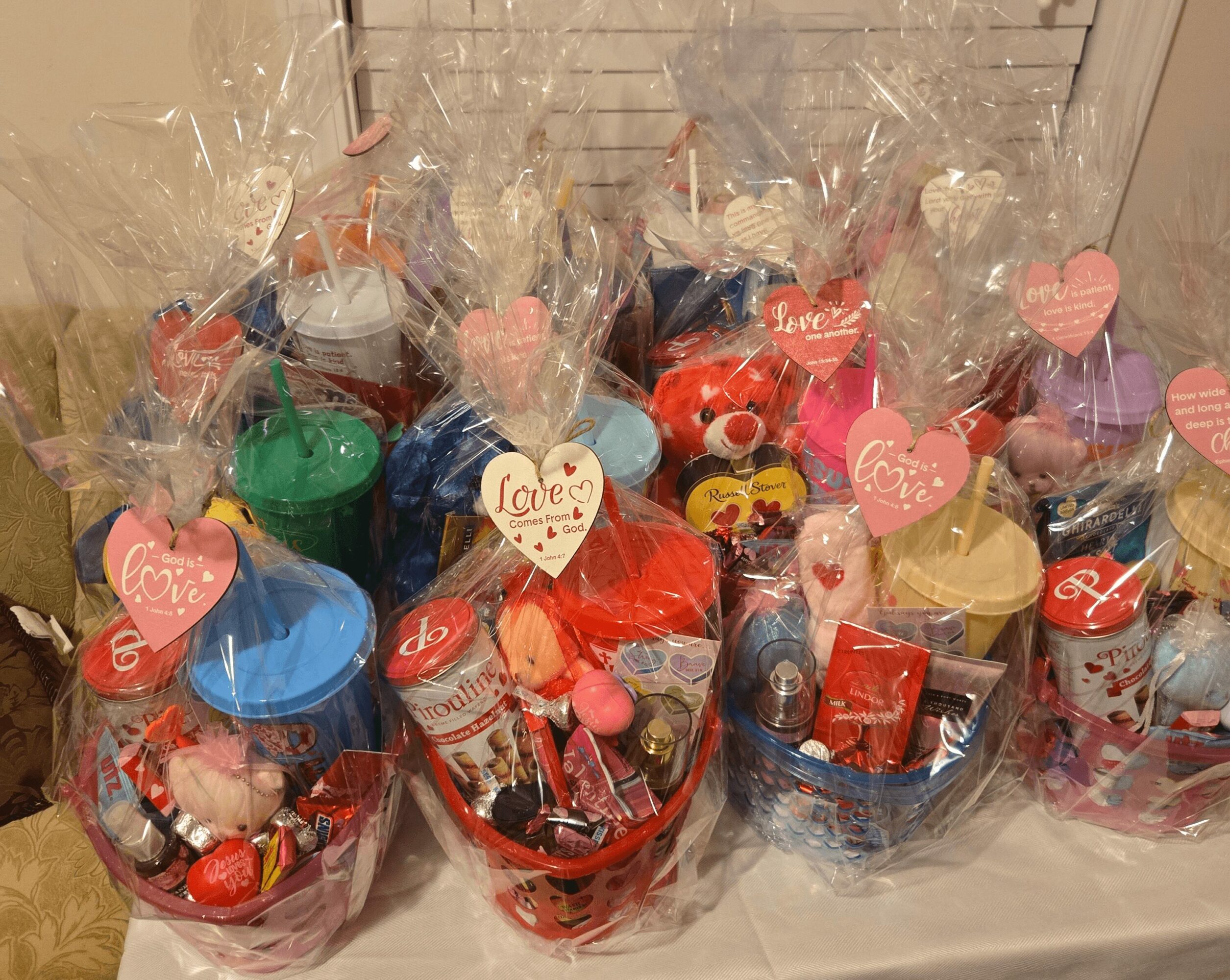 Multiple gift baskets wrapped in clear plastic with colorful items and pink heart-shaped tags, arranged on a table.