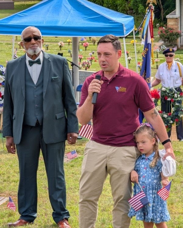 Three people standing outdoors at a memorial event, with a blue canopy and American flags in the background. One man is speaking into a microphone, a young girl holds flags, and another man stands beside them. The scene is on a grassy area with flowers and trees.