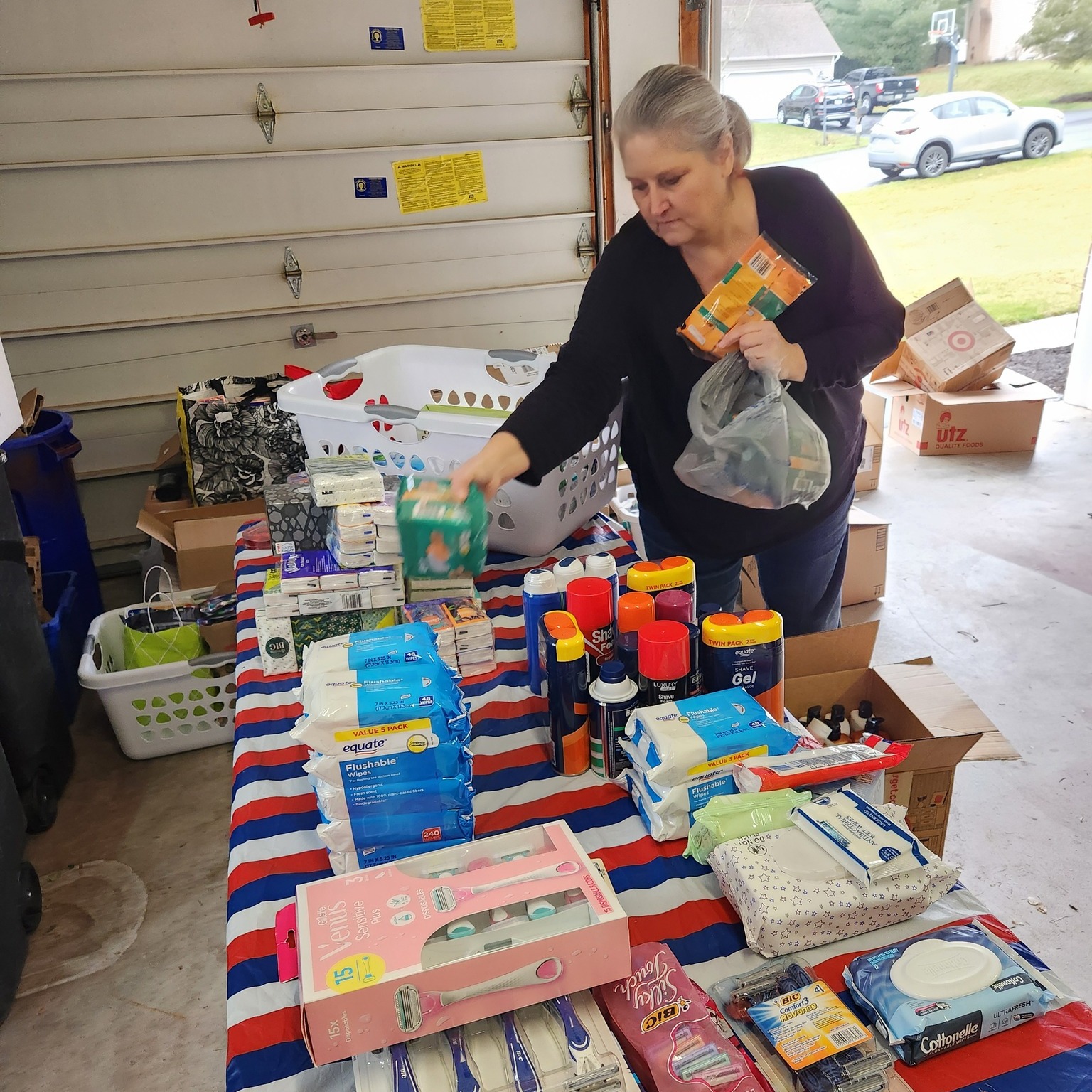 Woman shopping for household items at a table with cleaning supplies and paper products, inside a garage or storage area.