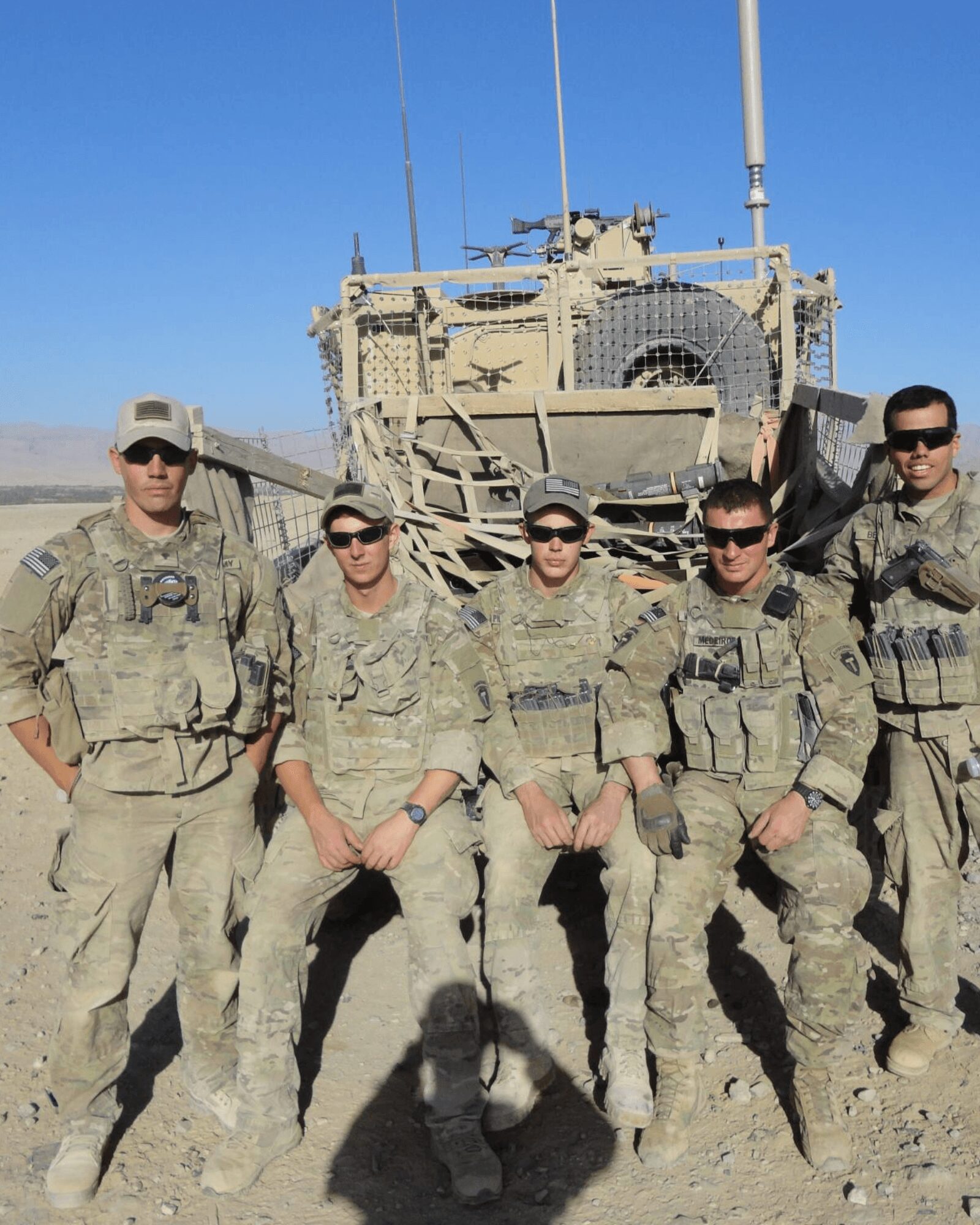 Six soldiers in uniform stand in front of a military vehicle on a dirt ground under a clear blue sky.