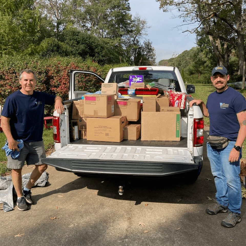 Two men stand beside a pickup truck filled with boxes outdoors, trees and a clear sky in the background.
