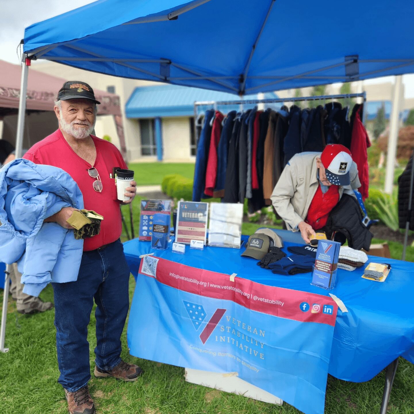 Man with gray beard in red shirt holding a coffee cup at outdoor booth with clothing rack and table, under blue canopy.