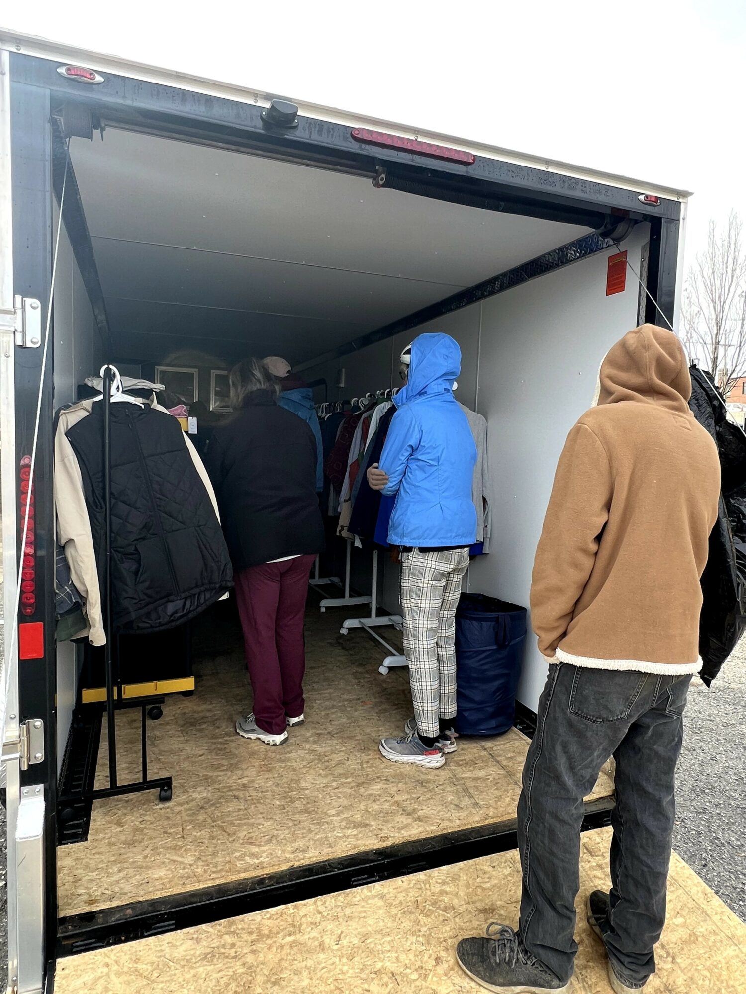 People inside a truck with clothing racks, some wearing hoodies and jackets, outdoors on a cloudy day.