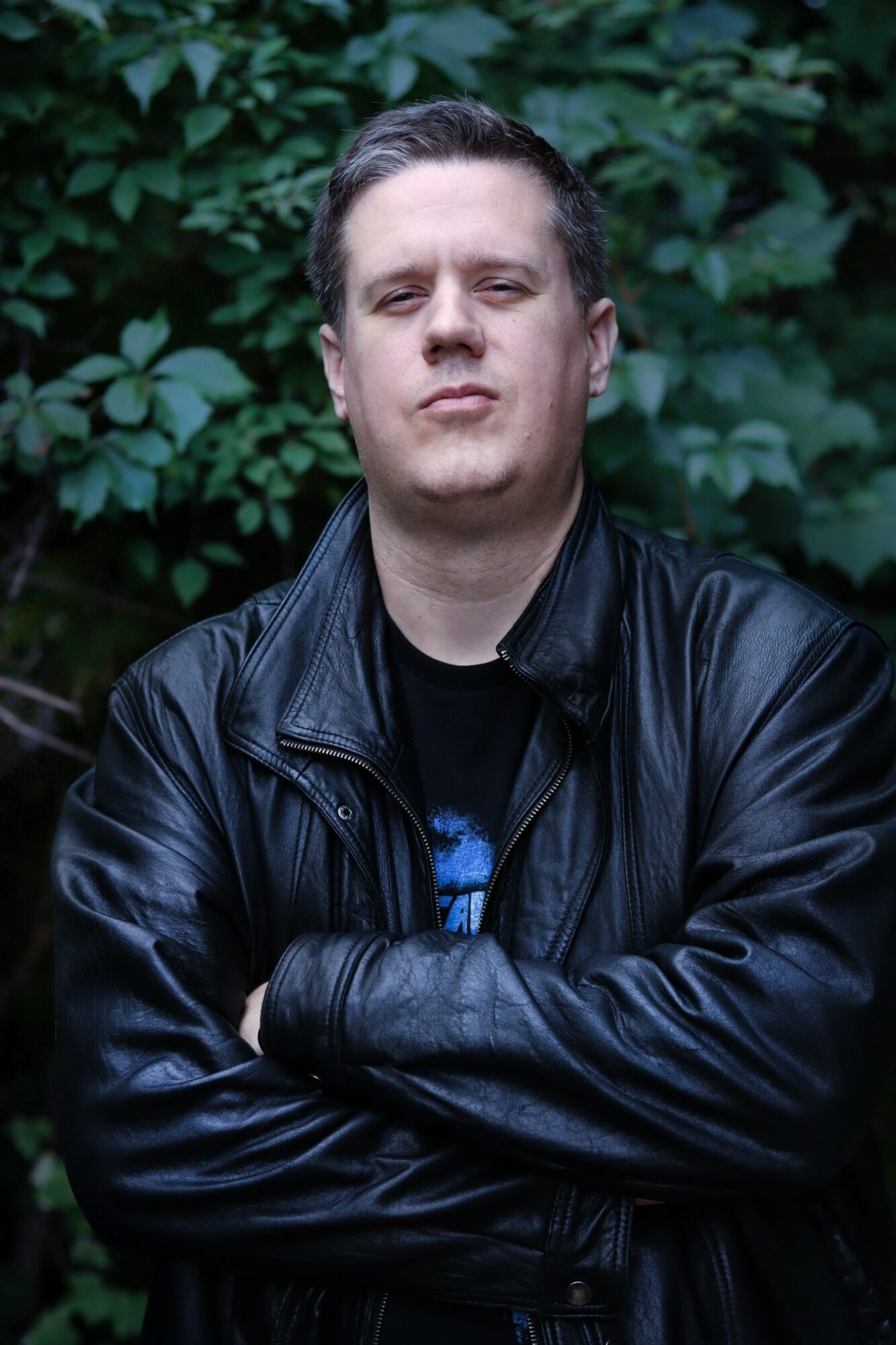 Man with short dark hair wearing a black leather jacket crosses arms in front of green foliage.