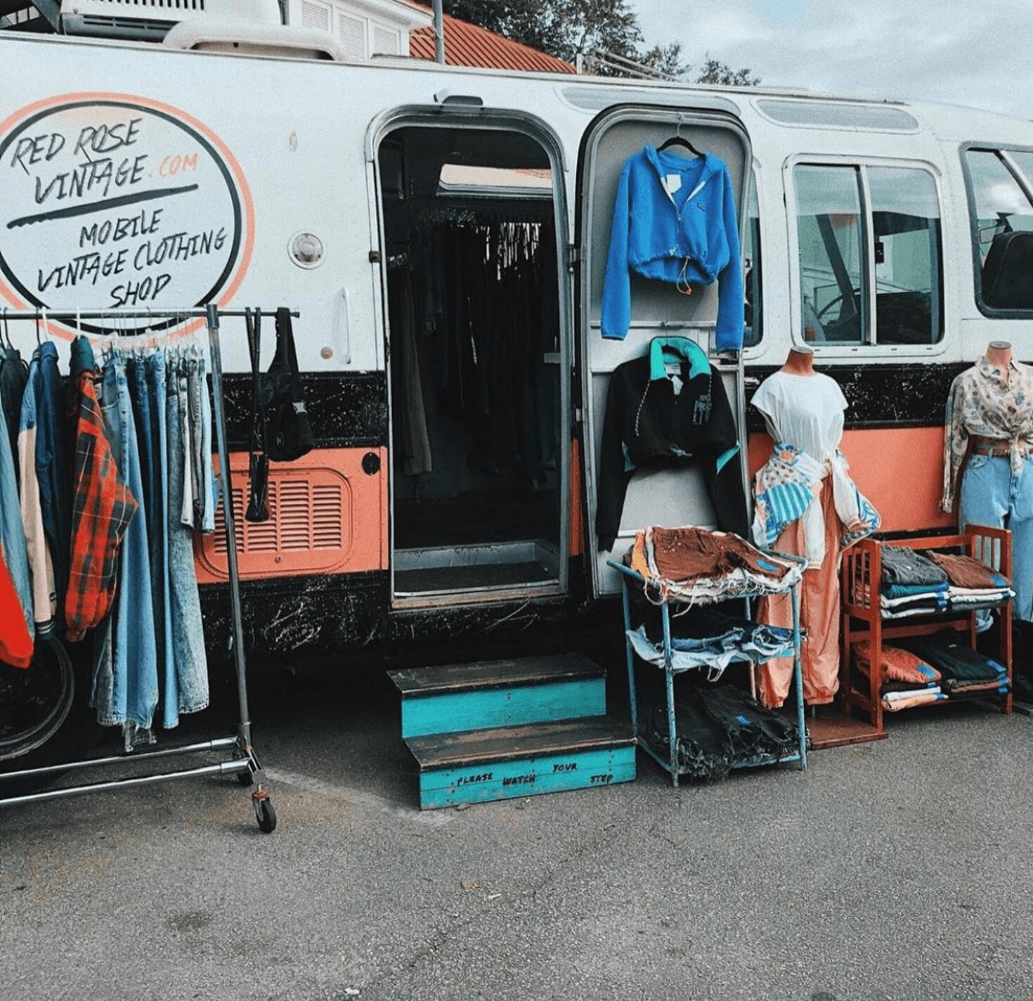 Vintage clothing shop set up outside a bus with racks of clothes and mannequins, sign reads 'Red Rose Vintage' and 'Mobile Vintage Clothing Shop'.