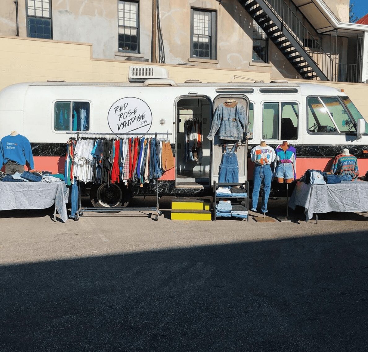 Outdoor clothing and accessories displayed on tables and racks in front of a vintage bus, with people browsing and a building in background.