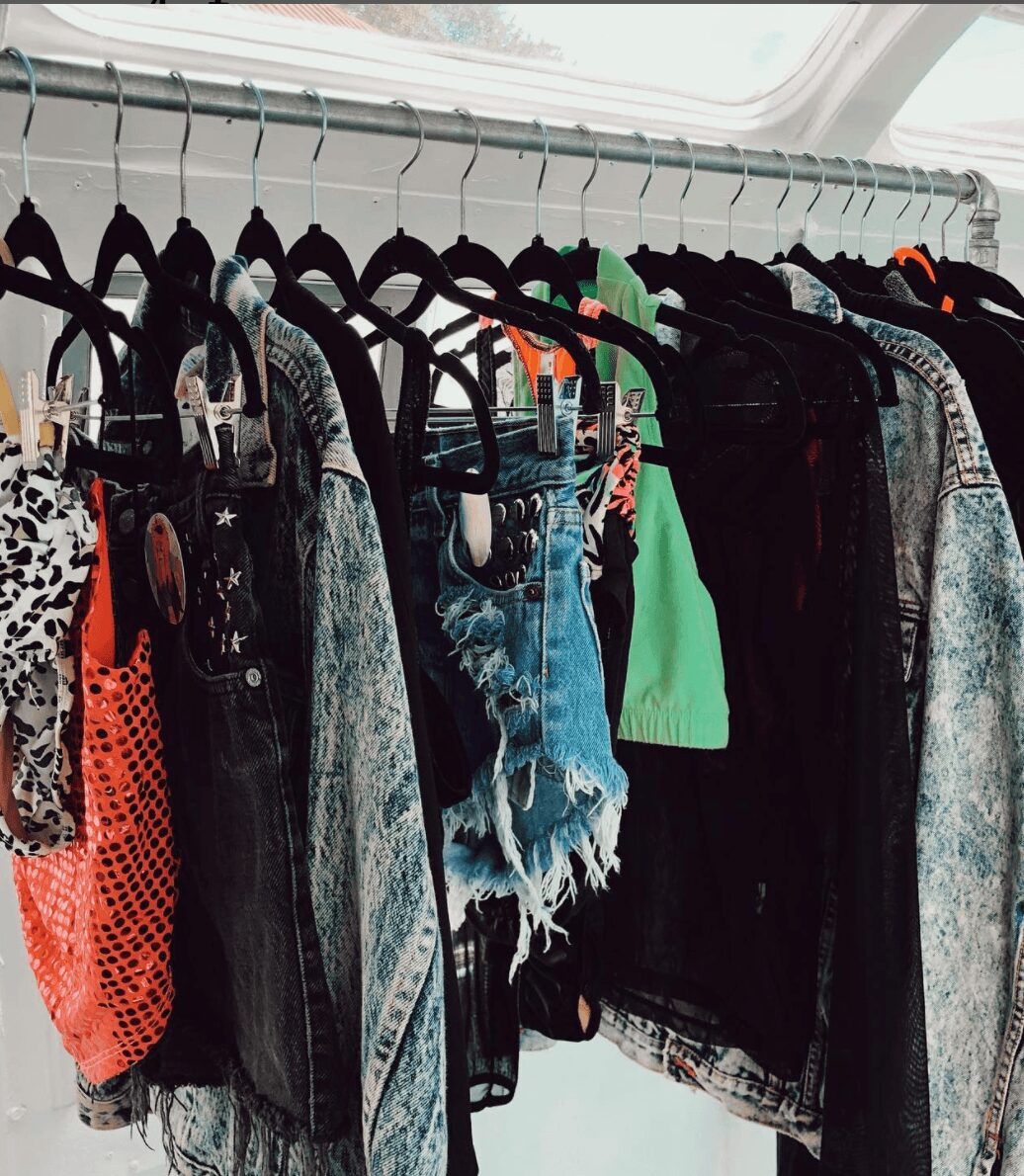 Clothes hanging on a rack inside a vehicle, including denim, black, and colorful garments, with a window in the background.