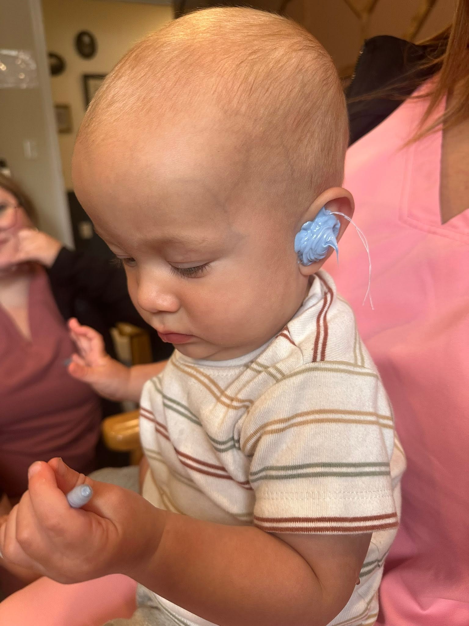 Young child with a shaved head and hearing aid, looking down, held by an adult in a pink shirt, indoors.