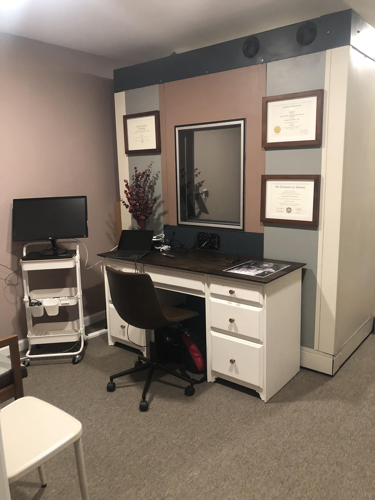 Desk with a chair, certificates on the wall, a mirror, and a small monitor on a cart in an office setting.