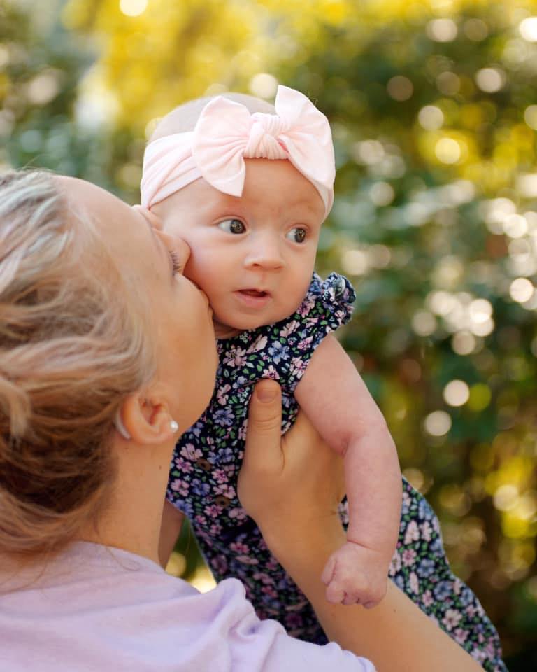 Adult holding a baby girl outdoors, baby wearing a pink headband with a bow, background of trees and sunlight.