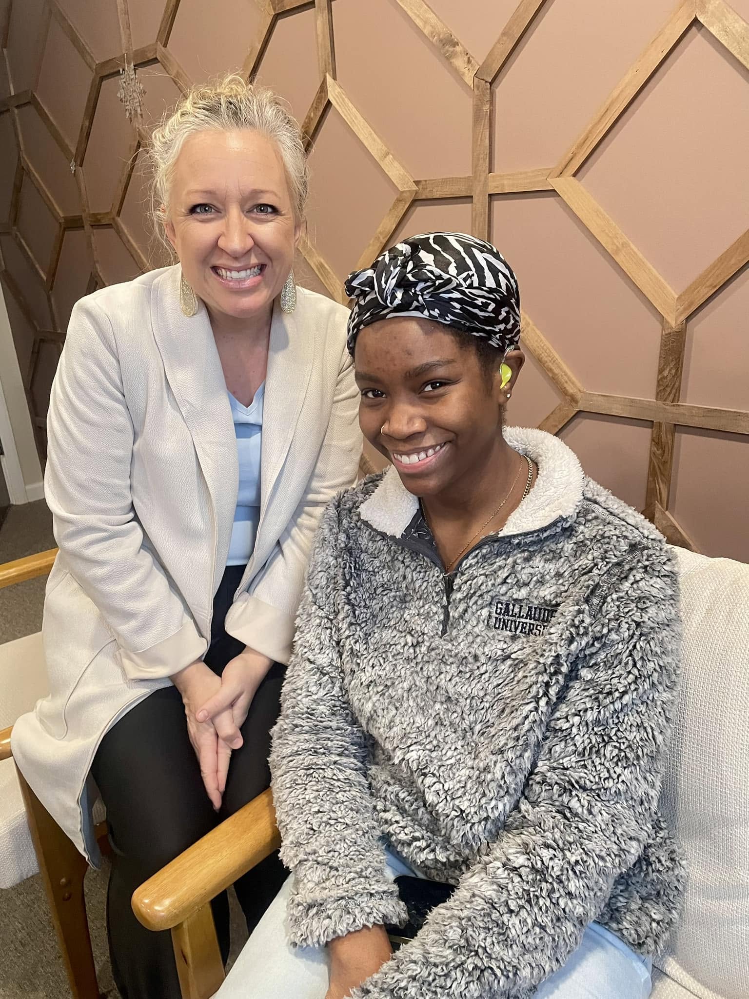 Two women smiling, one with light hair in a white blazer, the other with dark hair and a patterned headscarf, sitting indoors.