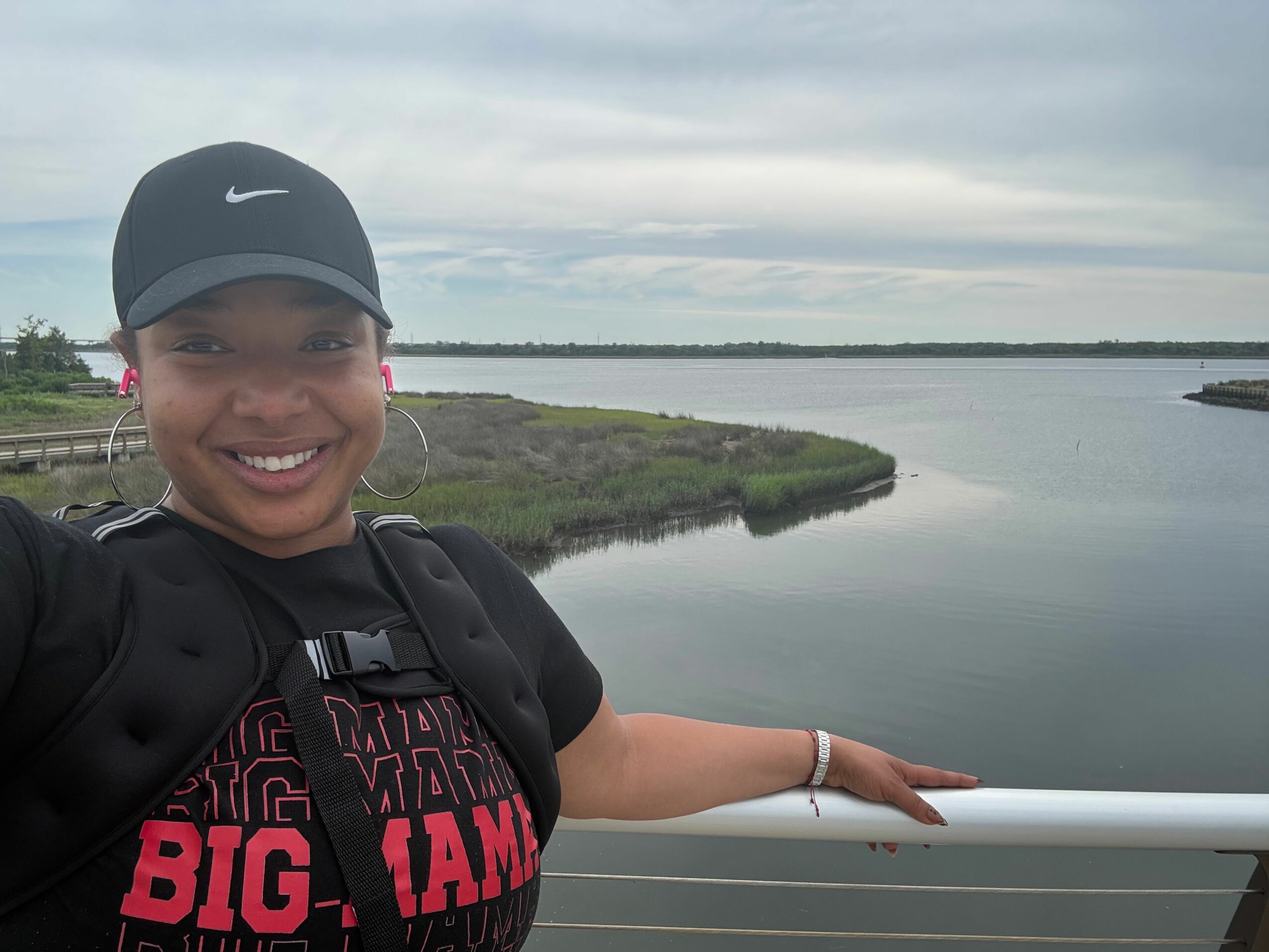 Person smiling, wearing a cap and black jacket, standing outdoors near water with a landscape in the background.