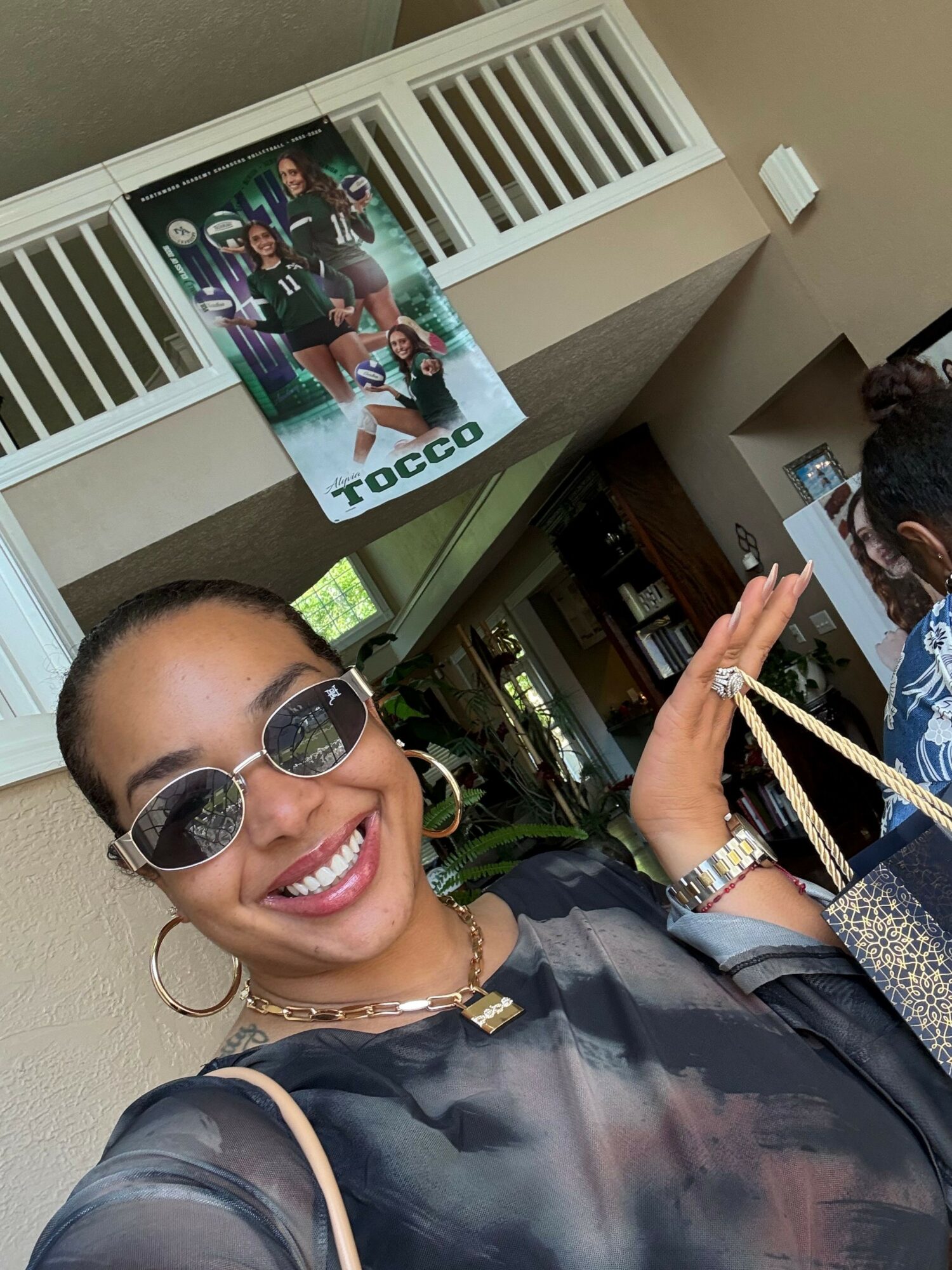 Person with short hair, sunglasses, and hoop earrings smiling, holding a drink, with a group photo on a table nearby.