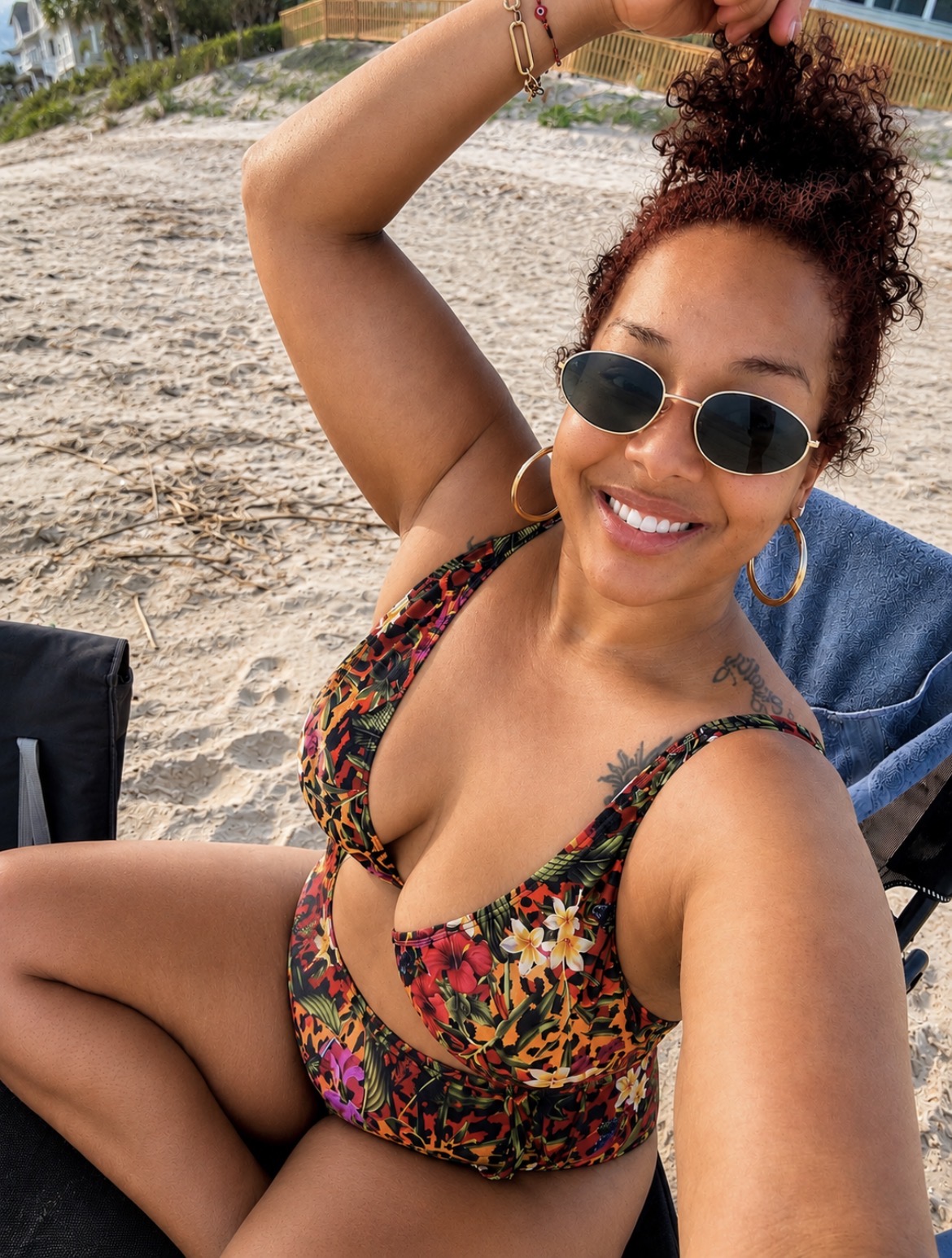 Smiling woman with curly hair and sunglasses at the beach, wearing a colorful swimsuit, sitting on a chair.