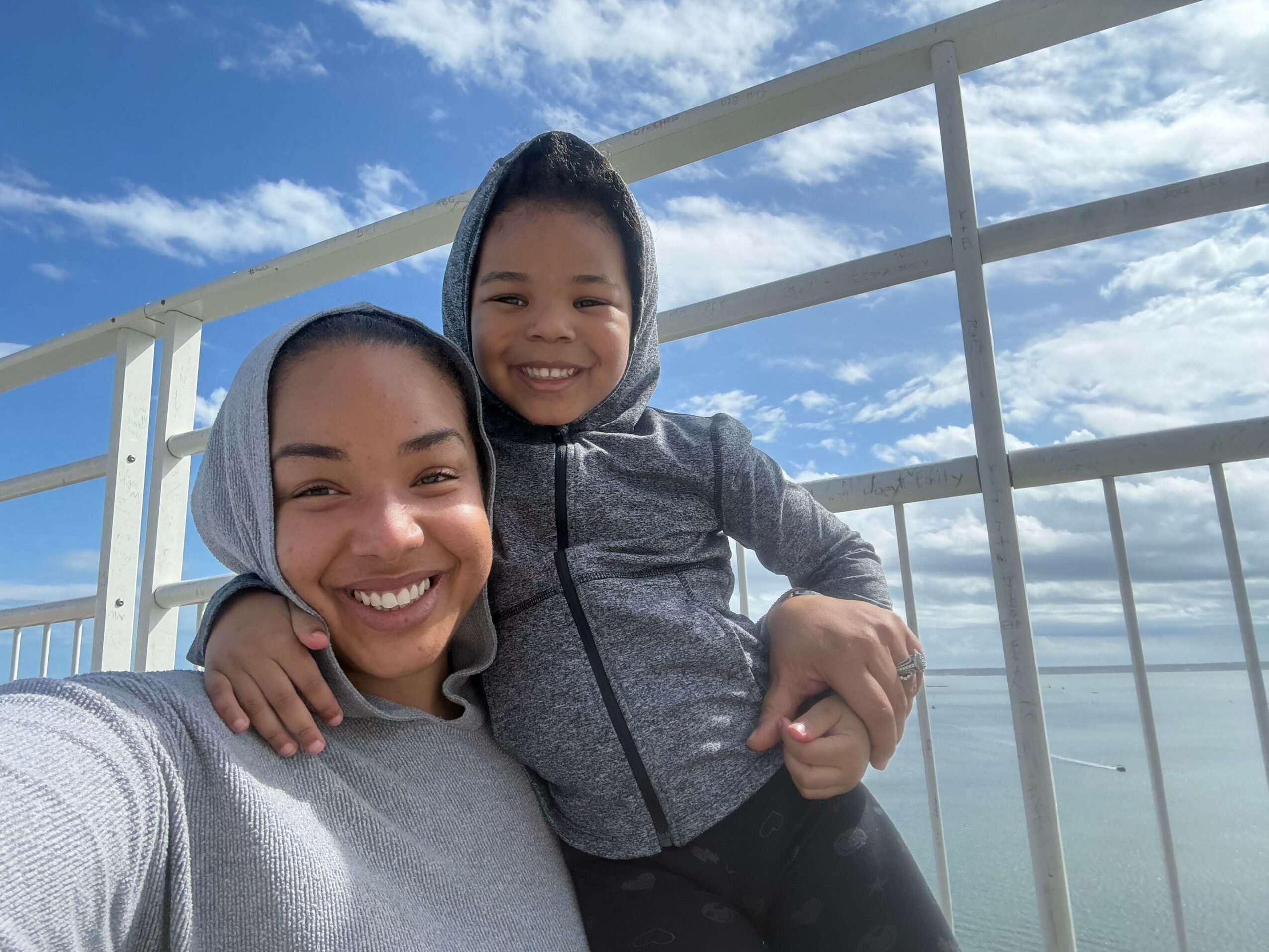 Two smiling women taking a selfie on a balcony with a blue sky and clouds in the background.