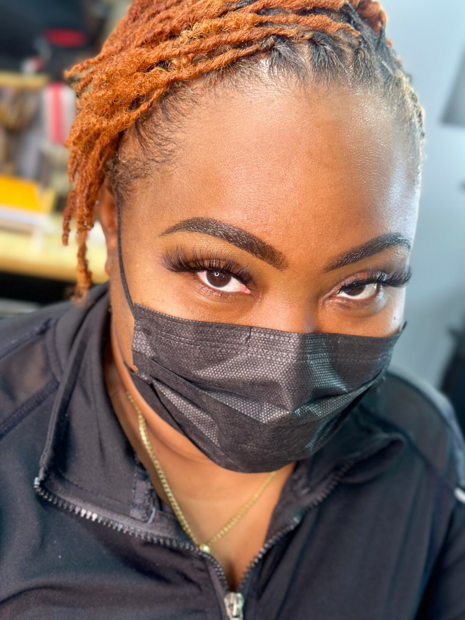 Close-up of a woman with styled orange dreadlocks, wearing a black face mask and dark clothing, looking at the camera.