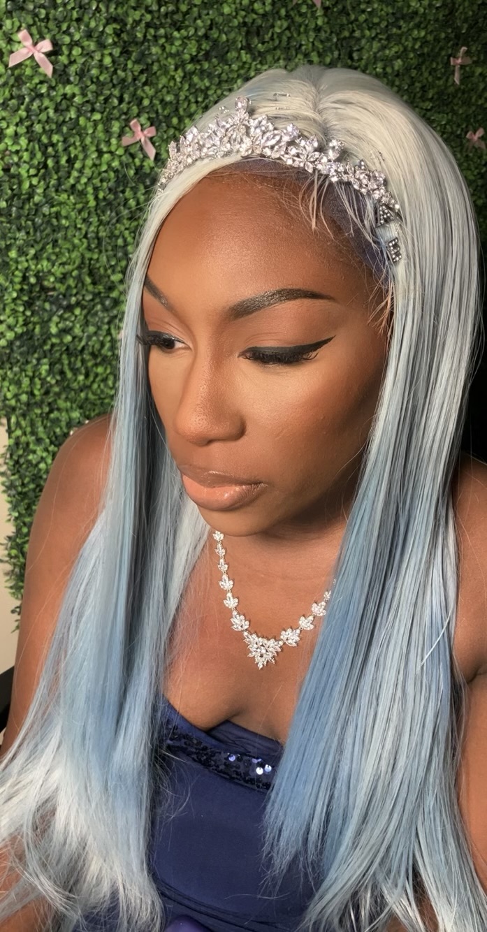 Woman with long, silver-blue hair wearing a tiara and necklace, standing against a green leafy background.