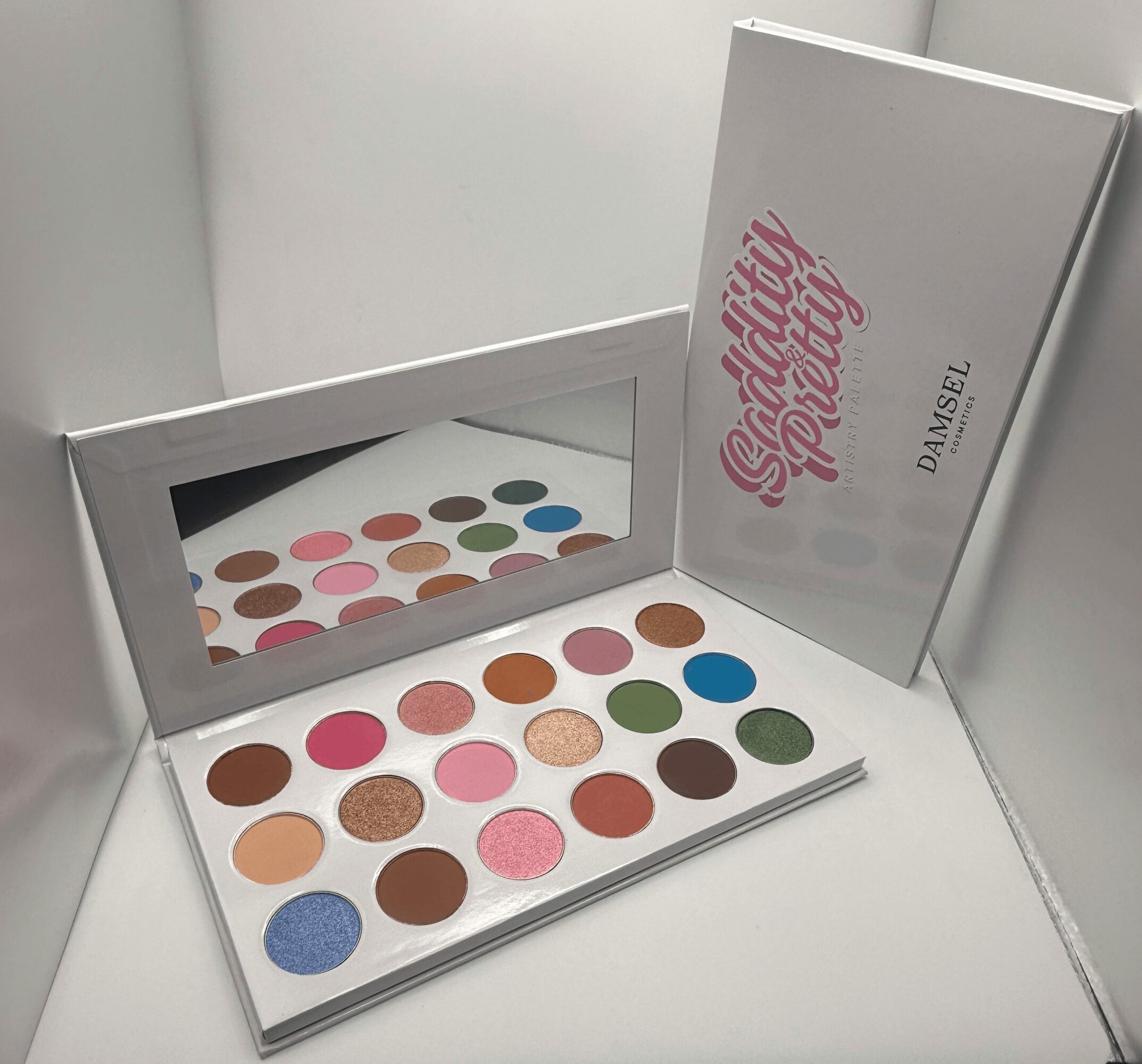 Open eyeshadow palette with 24 colorful shades, mirror, and a white box with pink text, on a white surface.