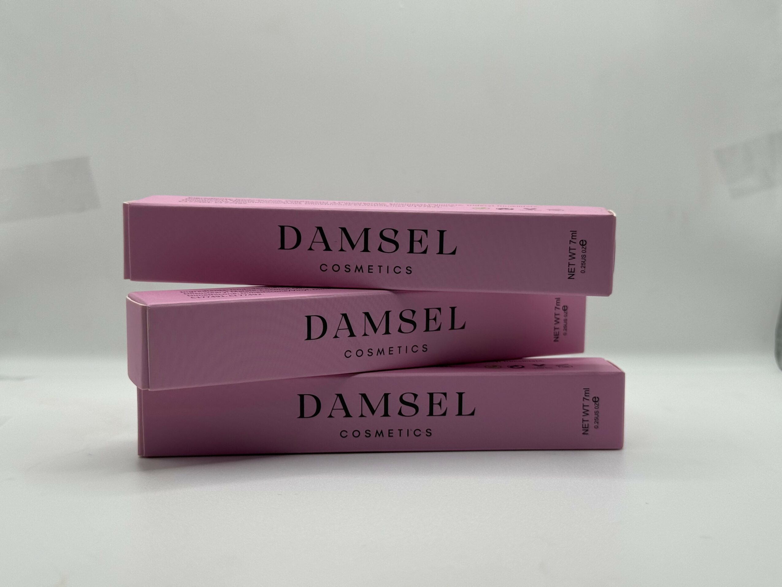 Three pink rectangular boxes labeled 'Damsel Cosmetics' stacked horizontally on a white surface.