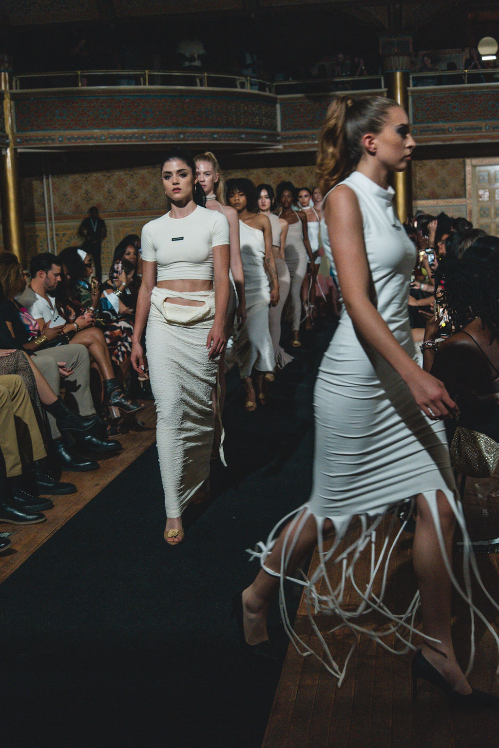 Models walk down runway in white outfits, audience seated on sides, ornate interior background.