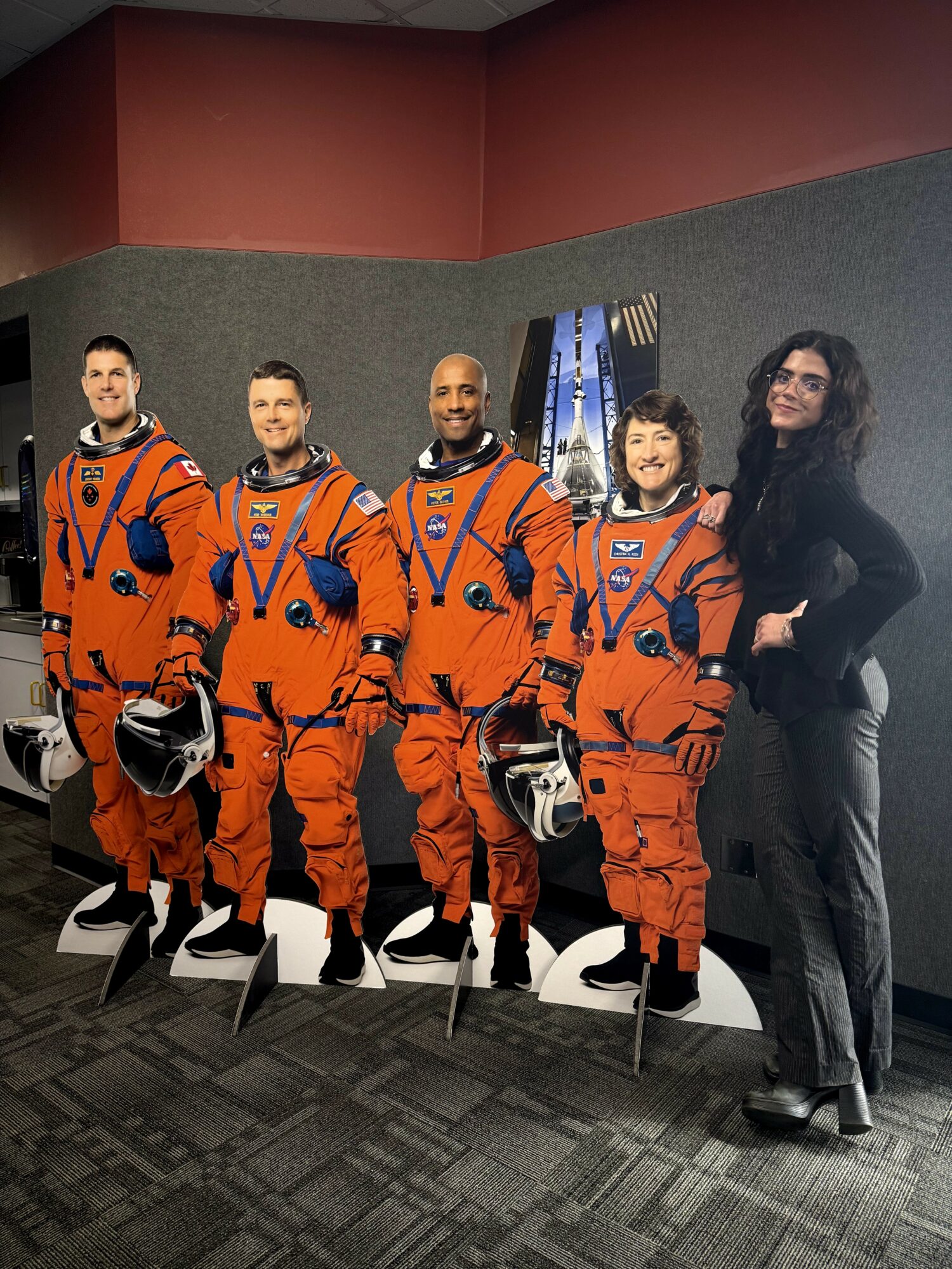 Five people, four in orange astronaut suits with helmets, stand in a row indoors, with a woman in black on the right.