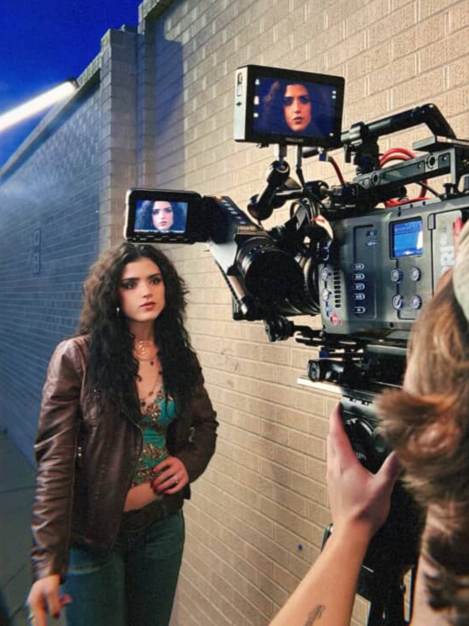 A woman with curly hair stands in front of a camera setup with monitors, against a brick wall background.