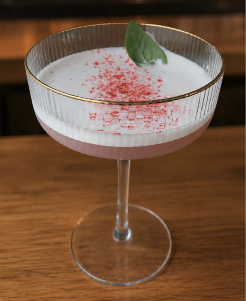 Cocktail in a glass with a green leaf garnish and red powder on top, on a wooden surface.