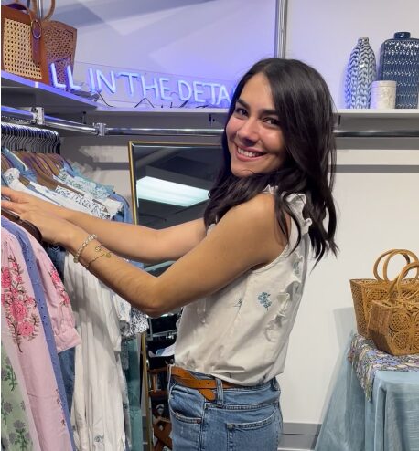 Woman shopping in clothing store, smiling, reaching for clothes on rack, wearing white top and jeans, store interior visible.