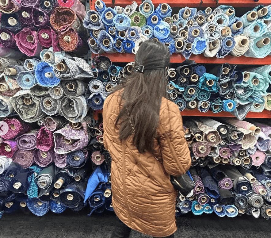 Person with long hair wearing a brown coat shopping for fabric rolls on shelves, facing away from the camera.