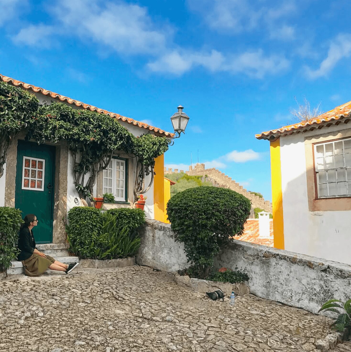 Person sitting on cobblestone street near colorful houses with bushes and a street lamp under a blue sky.