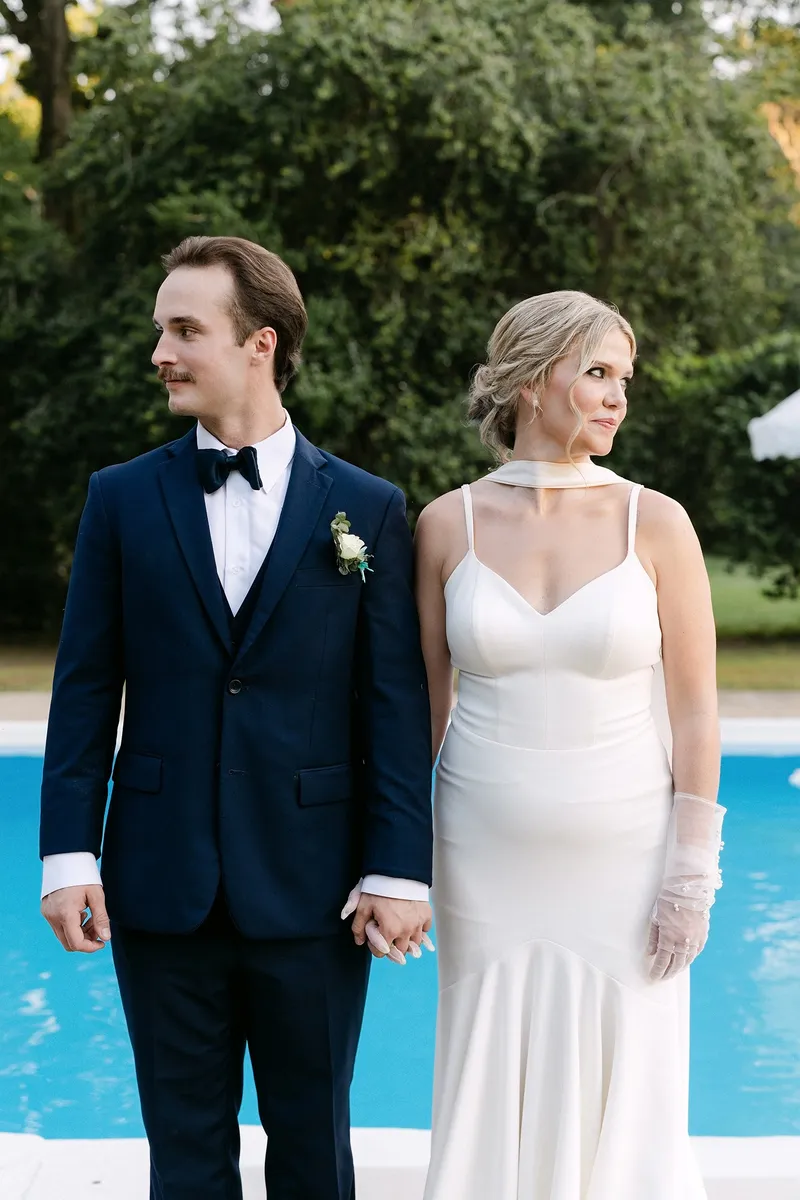 A couple in wedding attire holding hands outdoors near a pool, with trees in the background.