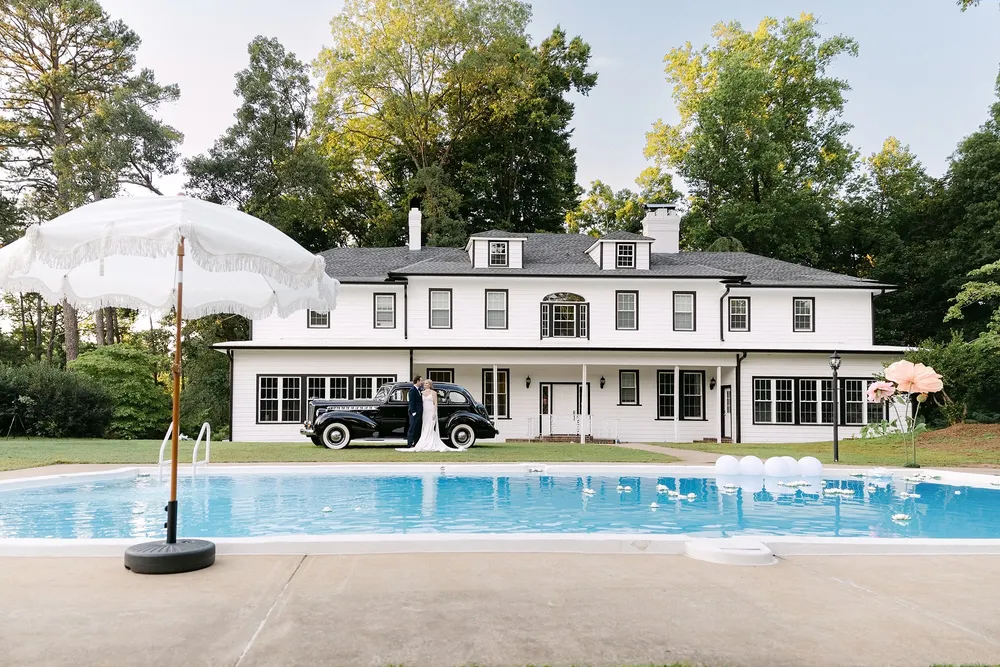 Large house with multiple windows and a garage, a black car parked in front, and a swimming pool with an umbrella nearby.