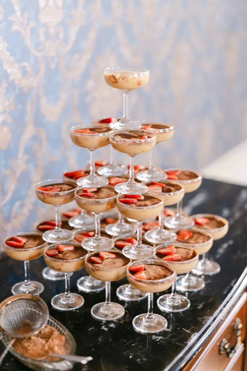A pyramid of champagne glasses filled with layered desserts topped with strawberries, set on a black surface against a blue patterned background.