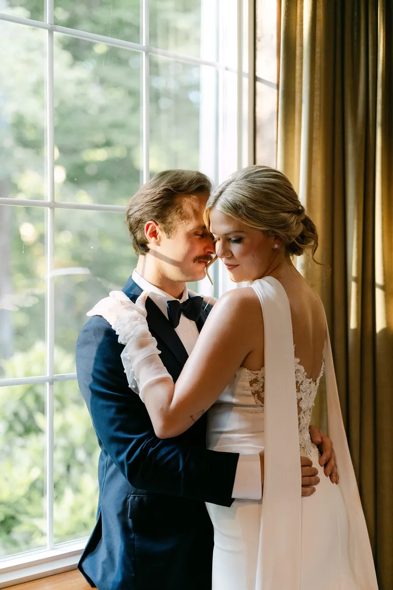 A couple in formal attire embracing indoors near a large window with curtains, sunlight streaming in, greenery outside.