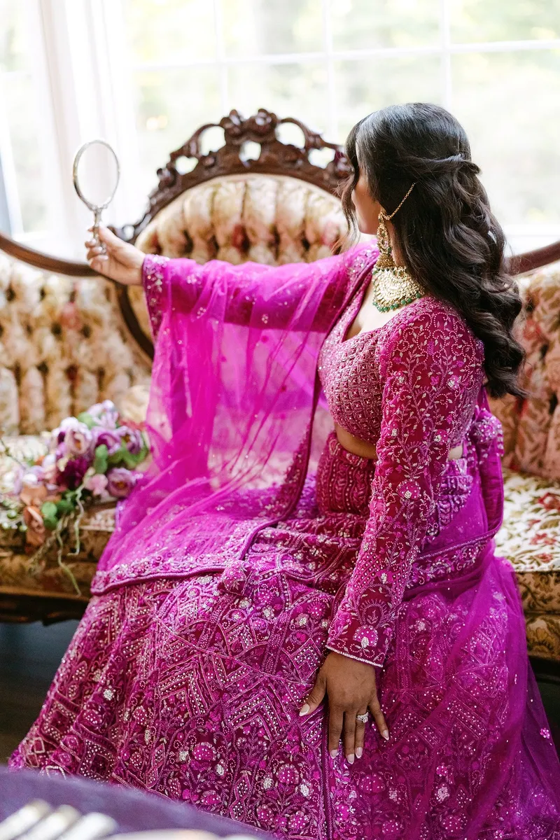 Woman in pink traditional dress holding a mirror, sitting on an ornate chair with flowers nearby.