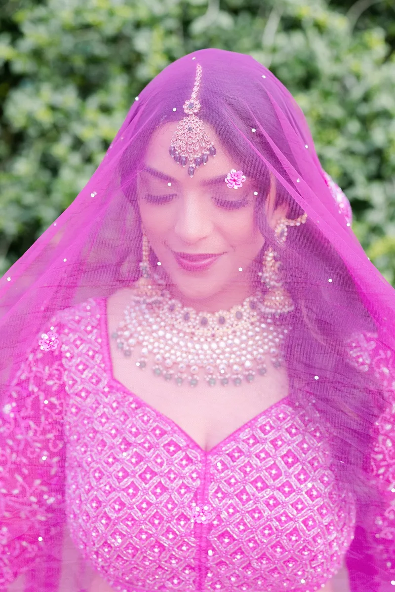Woman wearing pink traditional attire with jewelry, covered by a pink veil, outdoors with greenery in background.