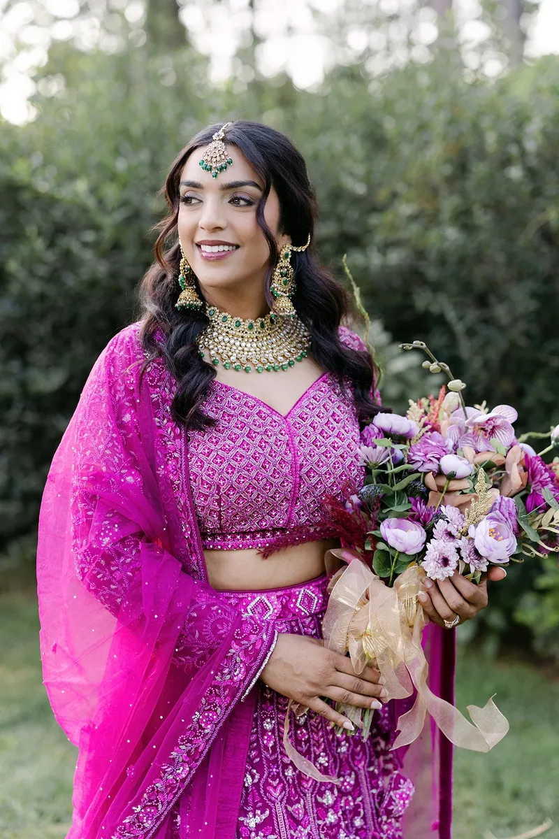 Woman in pink traditional attire holding a bouquet, outdoors with greenery background.
