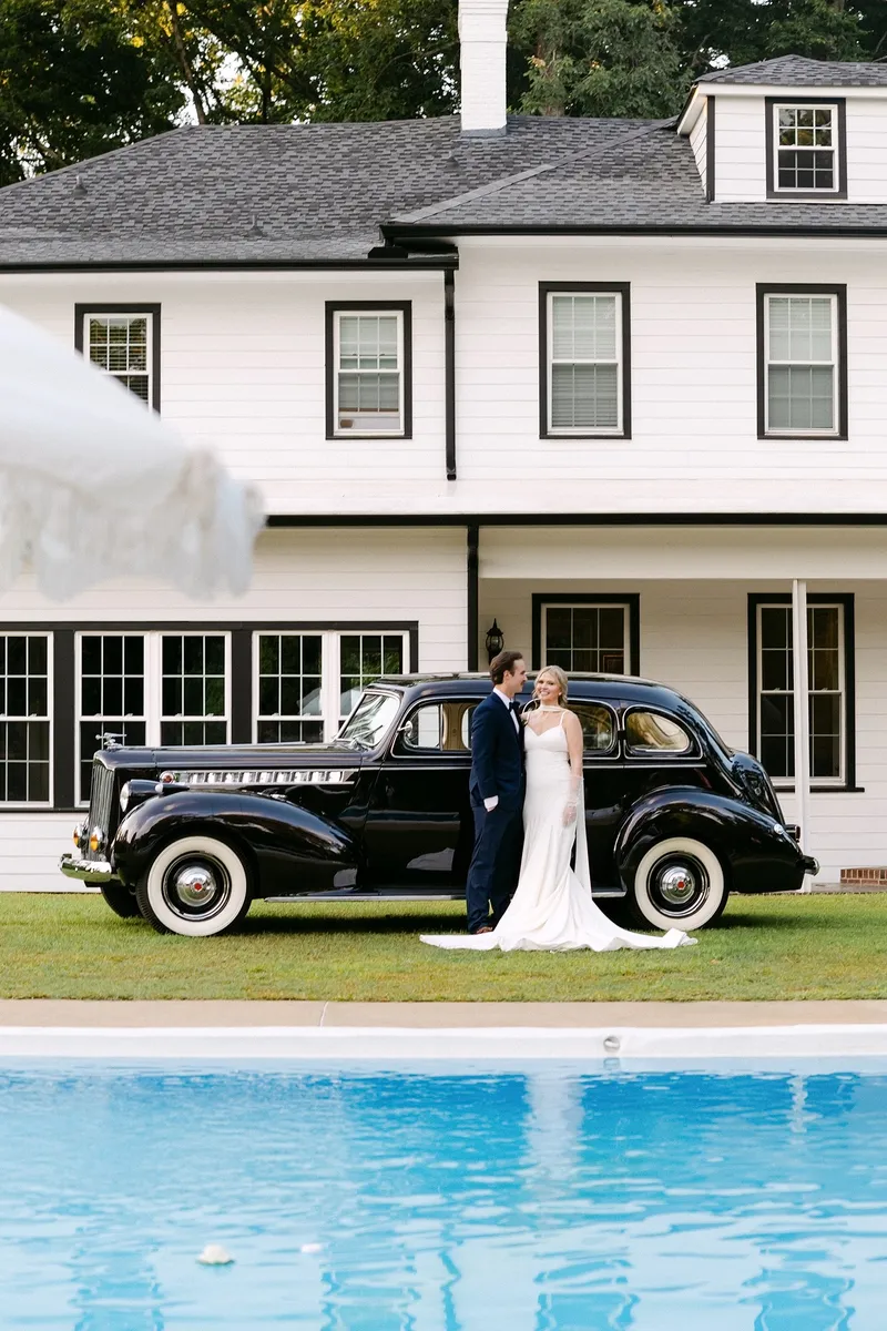 Couple in wedding attire stands beside a vintage black car in front of a white house with a pool in the foreground.