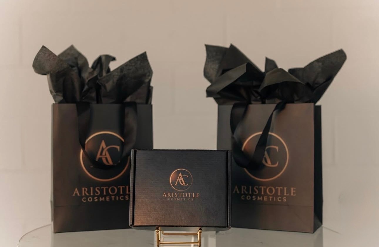 Two black shopping bags with black tissue paper and a black box with gold logo and text, displayed on a white surface.