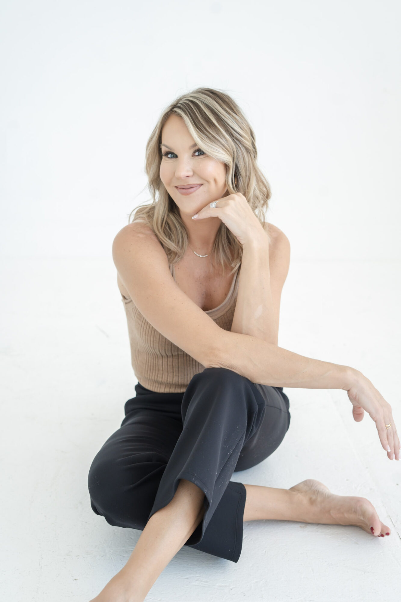 Woman with blonde hair sitting on the floor, smiling, wearing a beige top and black pants, against a white background.