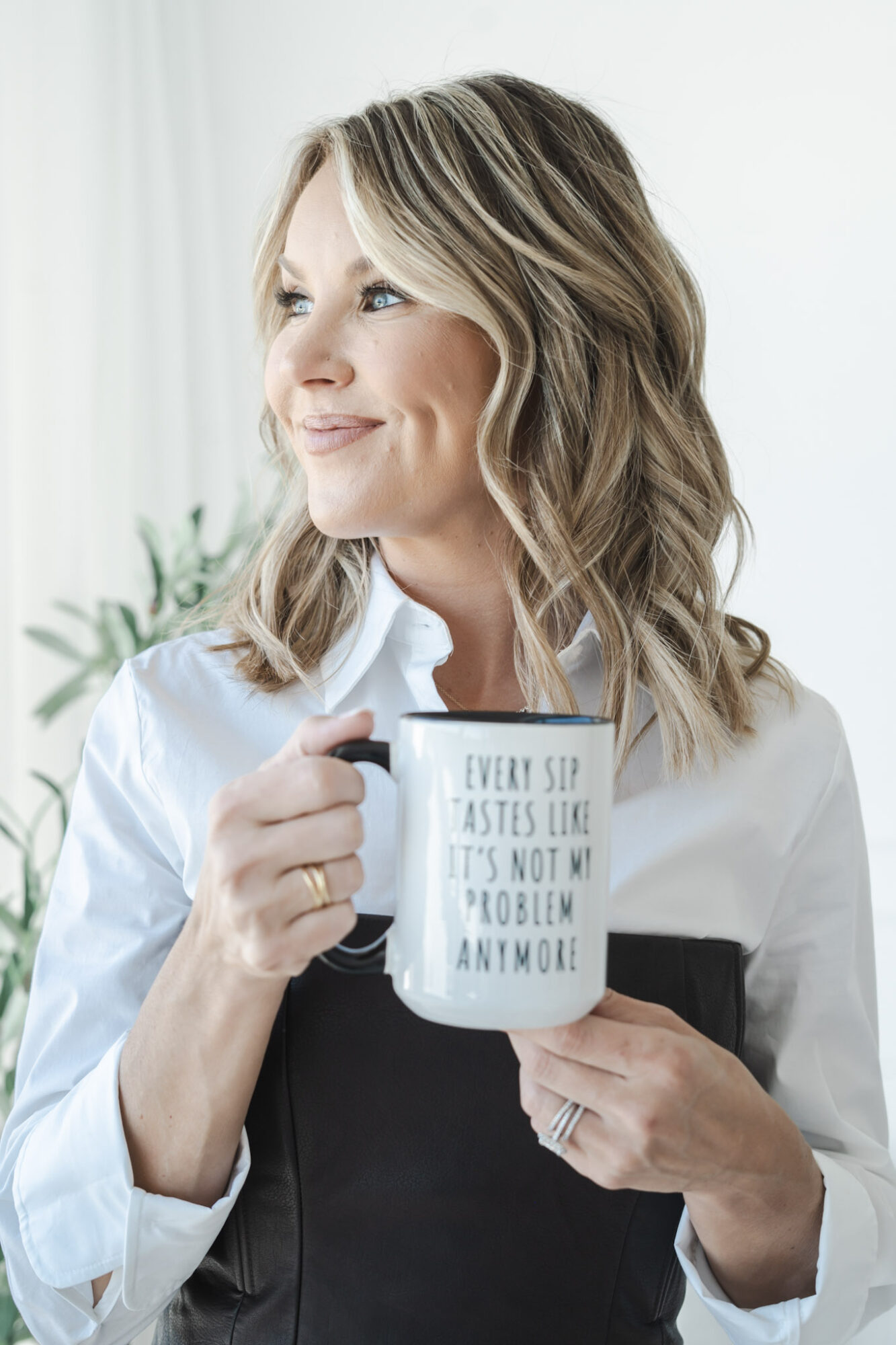 Woman with wavy hair holding a mug with text, smiling, in a bright room with a plant in the background.