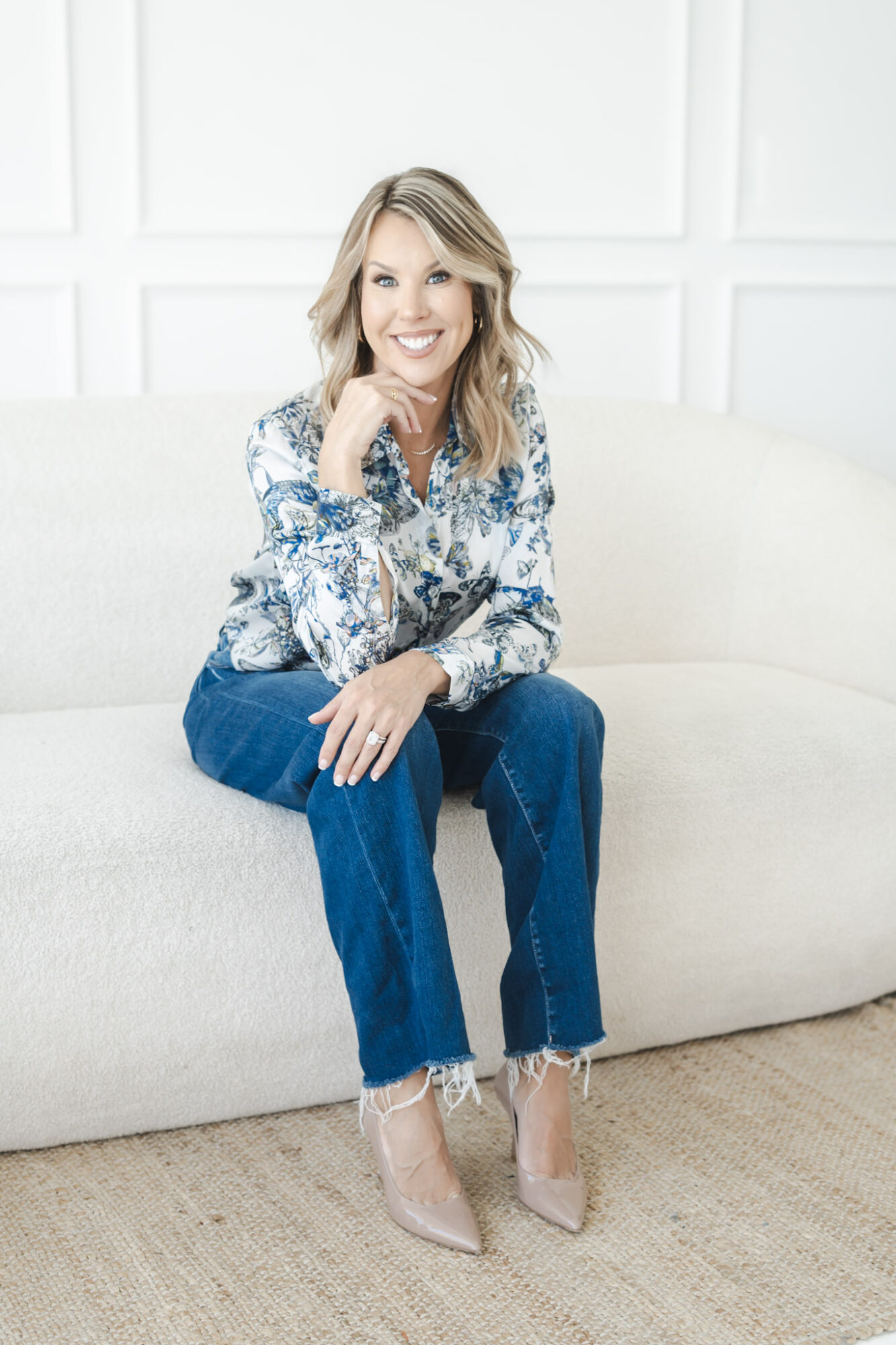 Woman sitting on a white sofa, smiling, wearing a patterned blouse, jeans, and high heels, in a bright room.