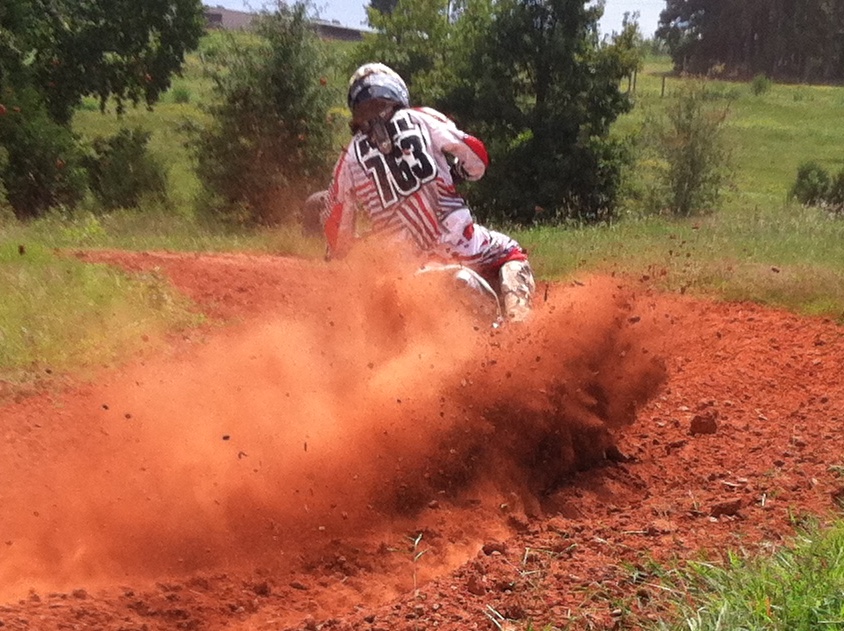 Motorcycle rider on dirt track kicking up dust, surrounded by green trees and grass, bright sunny day.