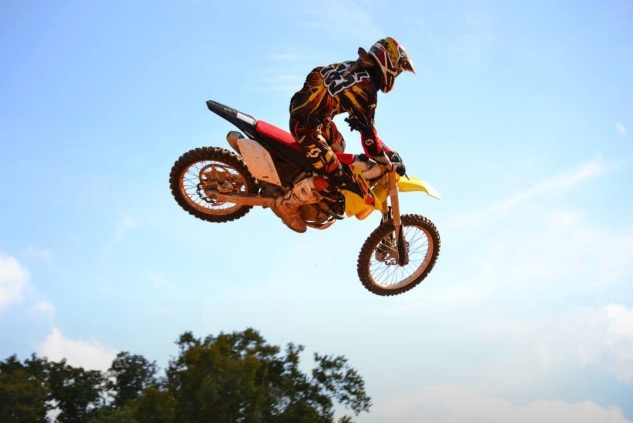 Motocross rider in mid-air against a blue sky, with trees below, performing a stunt on a yellow dirt bike.