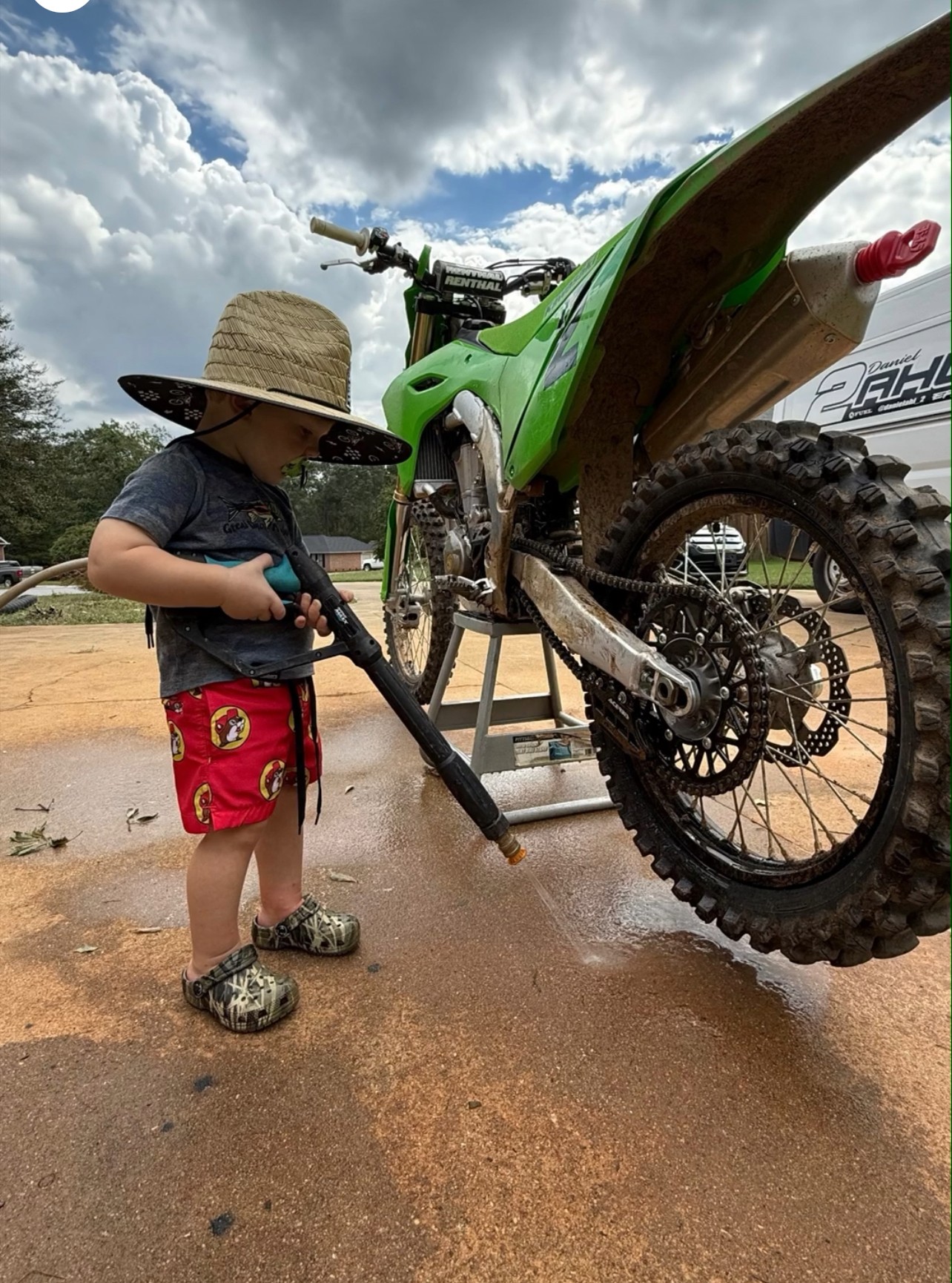 Child in hat and shorts cleaning a green dirt bike with a hose outdoors, cloudy sky in background.