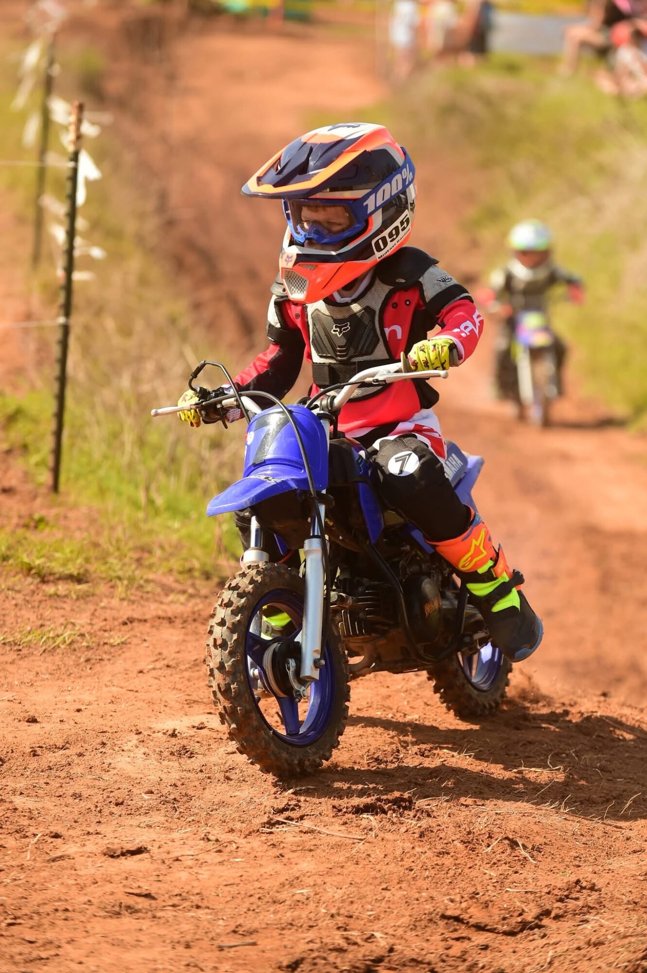 Child riding a blue dirt bike on a dirt trail, wearing a helmet and protective gear, with other riders in the background.